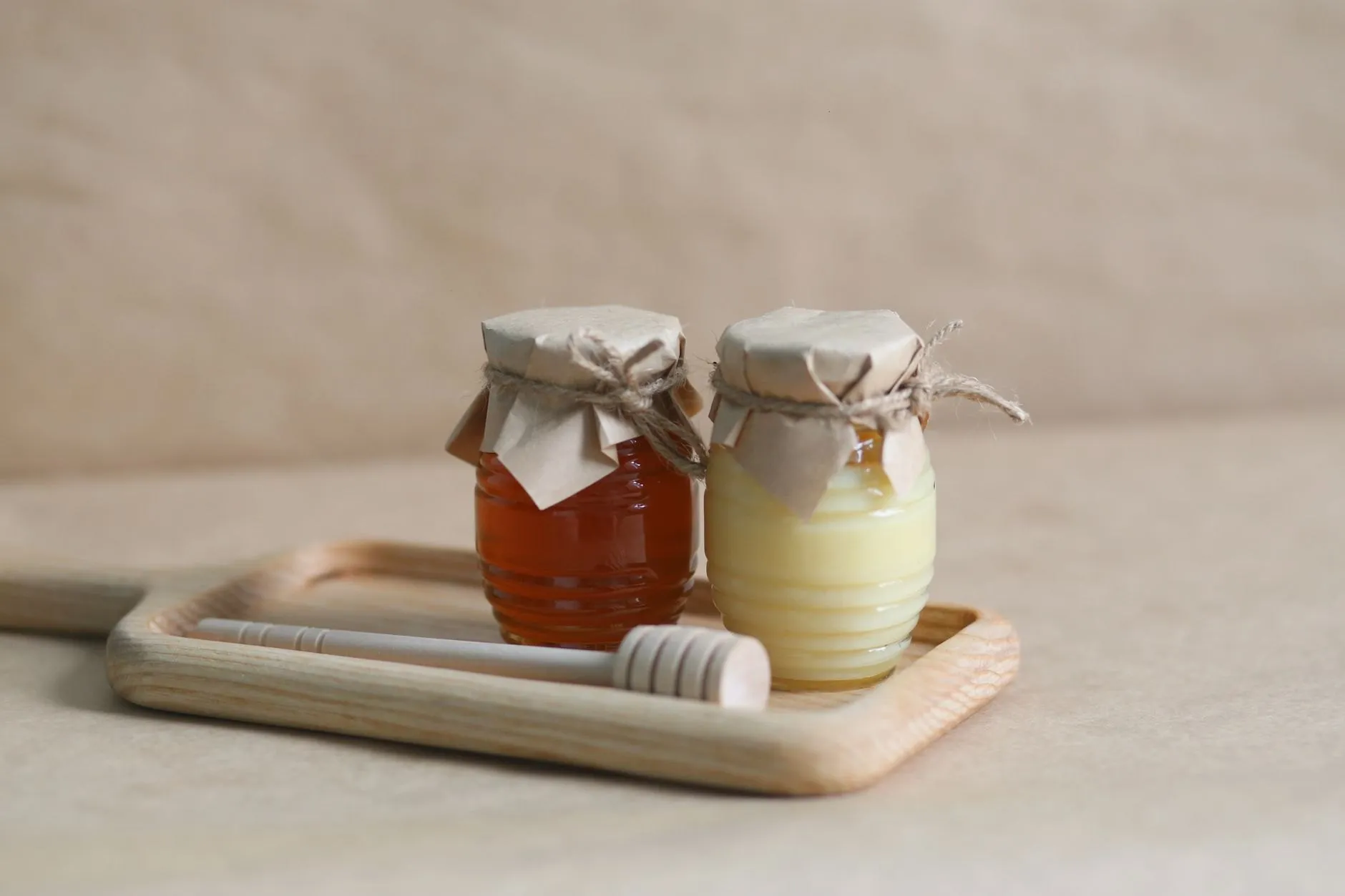 Two small honey jars with kraft paper tops on an oak tray with a dipper